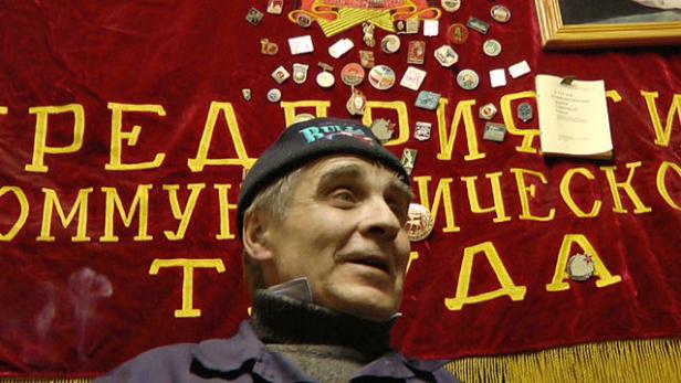 Ein Mann mit Mütze vor einer roten Fahne mit kyrillischer Schrift und Abzeichen.