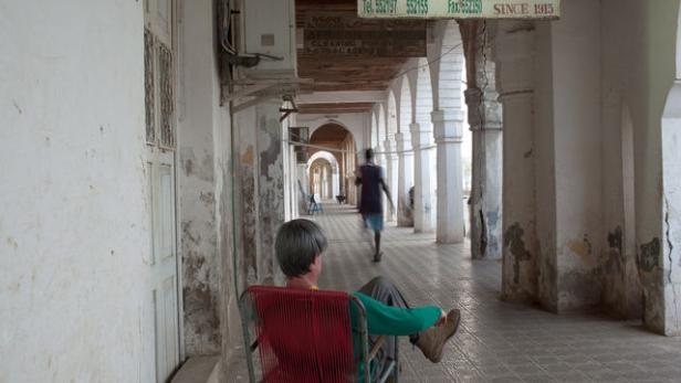 Ein Mann sitzt in einem roten Stuhl unter einem Säulengang, während eine Person vorbeigeht.