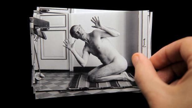 Eine Hand hält ein Schwarzweißfoto eines nackten Mannes, der auf einem Teppich kniet und die Hände hebt.