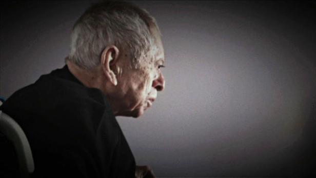 Ein älterer Mann sitzt in einem Rollstuhl und blickt nachdenklich zur Seite.