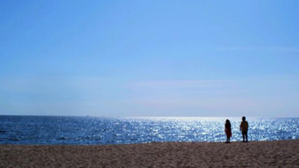 Zwei Personen stehen am Strand und blicken auf das glitzernde Meer.