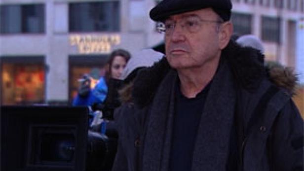 Ein älterer Mann mit Brille und Mütze steht vor einer Kamera, im Hintergrund ein Starbucks.