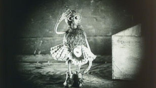 Eine anthropomorphe Figur mit Tutu und großen Augen steht vor einer Kiste.