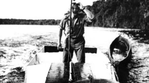 Ein Mann steht in einem kleinen Boot auf einem See und winkt.