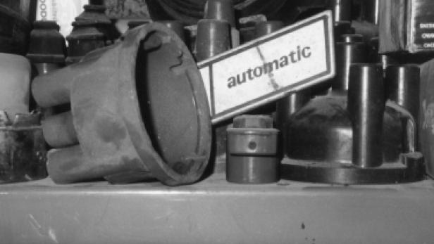 Verschiedene Autoteile liegen auf einem Regal, darunter ein Zündverteiler und ein Schild mit der Aufschrift „automatic“.
