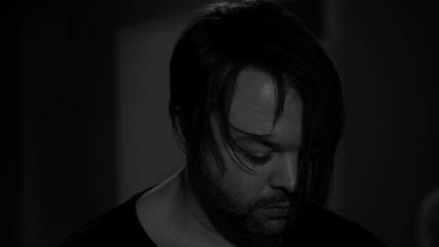 Ein Mann mit dunklen Haaren und Bart blickt nach unten.