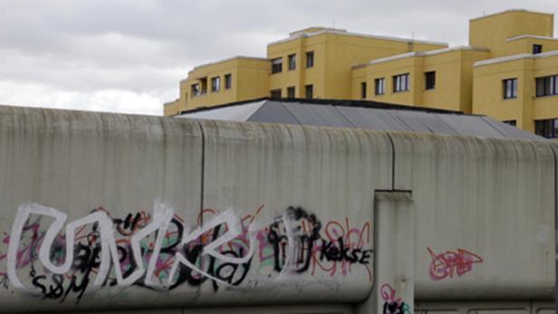 Eine Betonmauer mit Graffiti vor gelben Gebäuden unter einem grauen Himmel.