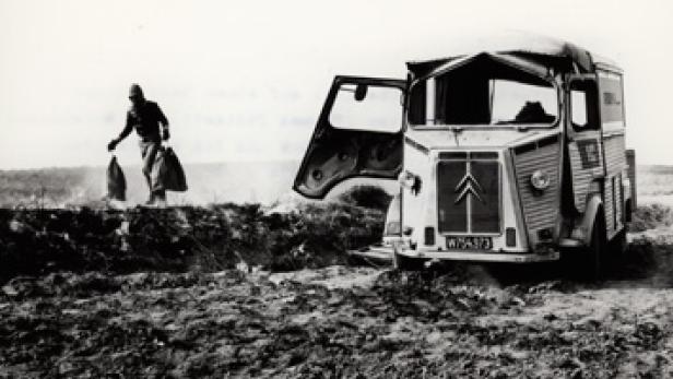 Ein Mann trägt Säcke an einem Strand, während ein Citroën-Lieferwagen im Sand steckt.