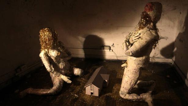 Zwei skulpturale Figuren mit goldenem Lametta knien vor einem kleinen Hausmodell in einem dunklen Raum.