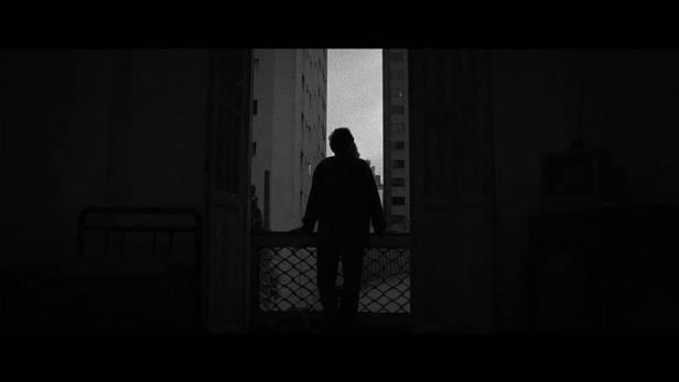 Eine Person steht in einer dunklen Wohnung am Fenster und blickt auf die gegenüberliegenden Gebäude.