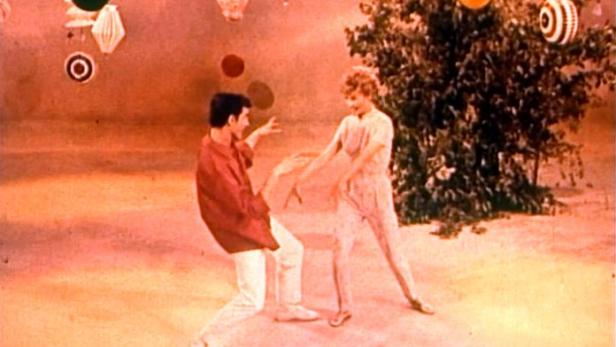 Ein Mann und eine Frau tanzen vor einem orangefarbenen Hintergrund mit Dekorationen.