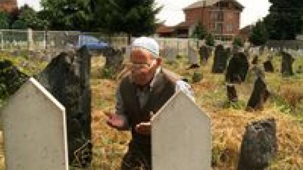 Ein älterer Mann mit Gebetskappe betet auf einem Friedhof vor zwei Grabsteinen.