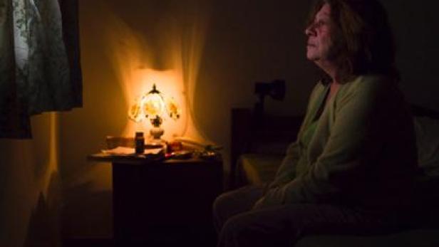 Eine Frau sitzt im Halbdunkel auf einem Bett und schaut nachdenklich zur Seite.