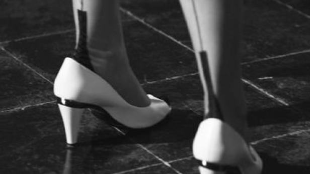 Nahaufnahme von Beinen in Strümpfen und weißen Schuhen mit Absatz auf einem gefliesten Boden.