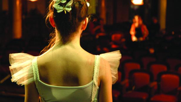 Eine Ballerina mit Schmetterlingen im Haar steht auf der Bühne vor leeren Sitzen.
