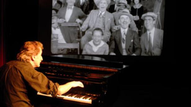 Ein Pianist spielt vor einer Leinwand, auf der eine Schwarzweißaufnahme von lachenden Menschen zu sehen ist.