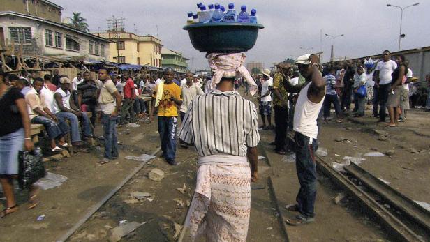 Eine Frau balanciert eine Schüssel mit Wasserflaschen auf dem Kopf in einer belebten Straße.