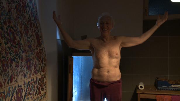 Ein älterer Mann steht mit erhobenen Armen in einem Badezimmer.