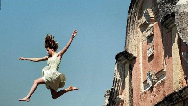 Eine junge Frau springt barfuß vor einem alten Gebäude in die Luft.