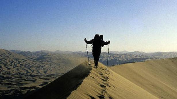 Ein Wanderer mit Rucksack und Stöcken geht über eine Sanddüne.