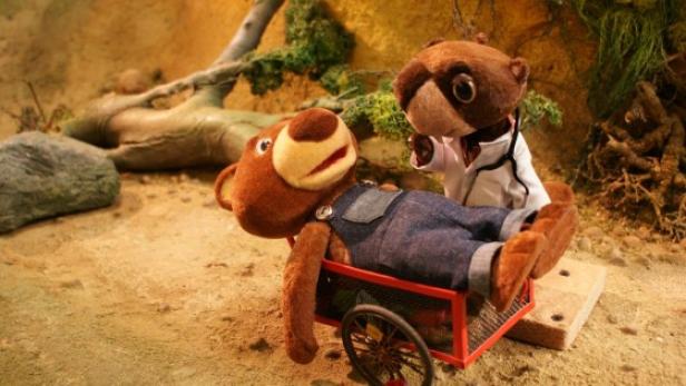 Ein Bär liegt in einem Wagen, während ein Dachs im Arztkittel daneben steht.