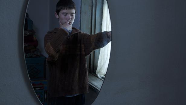 Ein junger Mann betrachtet sein Spiegelbild in einem ovalen Spiegel.