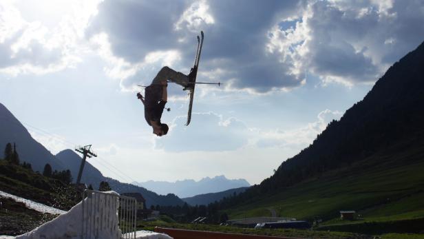 Ein Skifahrer macht einen Rückwärtssalto in der Luft vor einer Bergkulisse.