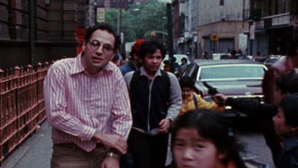 Ein Mann mit Brille und gestreiftem Hemd steht auf einem Bürgersteig in einer belebten Straße.