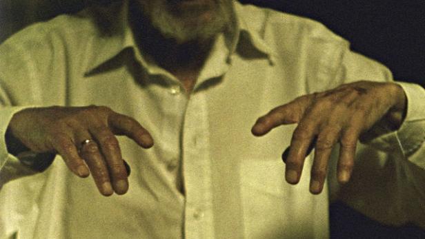 Ein älterer Mann mit Bart und weißem Hemd hält seine Hände vor sich.