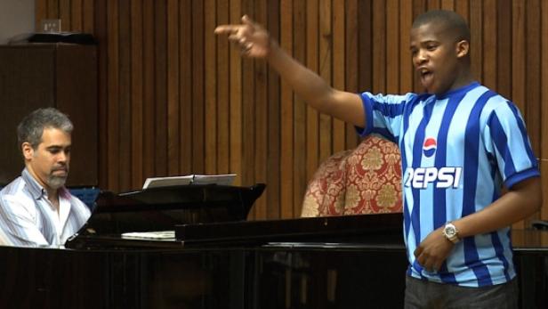 Ein junger Mann singt mit erhobenem Arm, während ein Mann Klavier spielt.