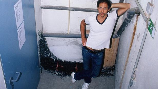 Ein Mann posiert lässig in einem Kellerraum mit Rohren und einer Tür.