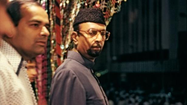 Zwei Männer, einer mit Brille und traditioneller Kopfbedeckung, stehen vor einer Menschenmenge.