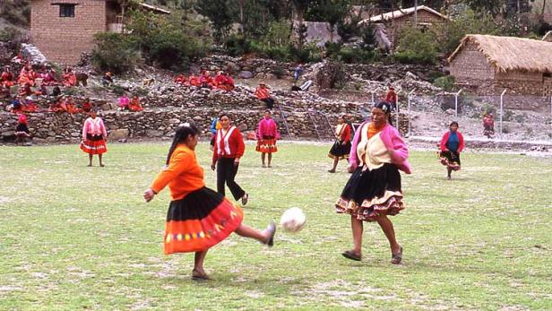 Frauen in traditioneller Kleidung spielen Fußball auf einem grünen Feld.