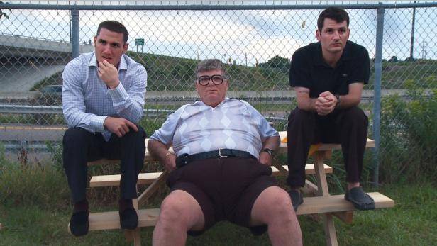 Drei Männer sitzen auf einer Picknickbank vor einem Zaun.