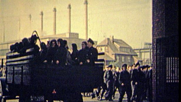 Ein Lastwagen transportiert Menschen in Uniform vor dem Hintergrund einer Industrieanlage.