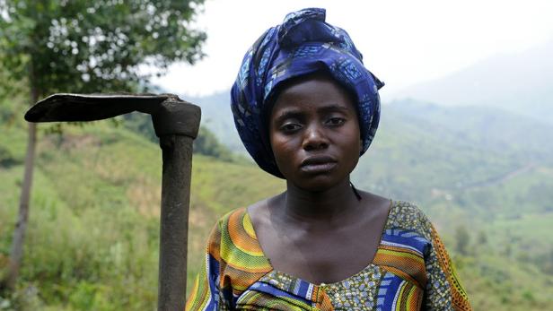 Eine Frau mit Kopftuch hält eine Hacke vor einer hügeligen Landschaft.