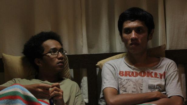 Zwei junge Männer sitzen auf einem Bett vor einem Fenster.