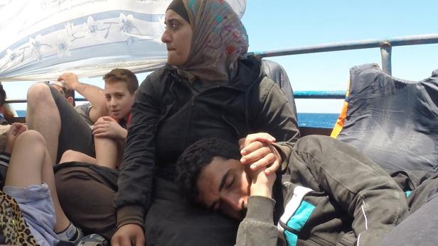 Eine Frau mit Kopftuch hält die Hand eines schlafenden Mannes auf einem Boot.