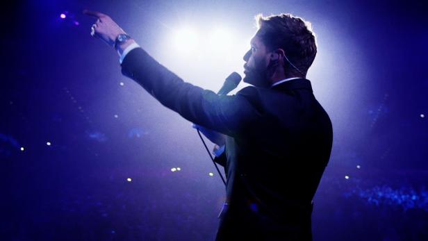 Ein Mann in einem Anzug steht auf einer Bühne und zeigt mit dem Finger nach oben.