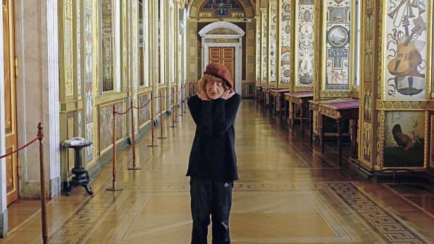 Eine ältere Frau mit Hut steht in einem reich verzierten Korridor.