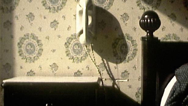 Ein altmodisches Telefon hängt an einer Wand mit Blumenmuster neben einem Nachttisch.