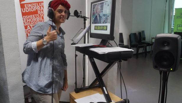 Eine Frau mit Kopfbedeckung und Kopfhörern steht vor einem Monitor in einem Studio und gibt einen Daumen hoch.