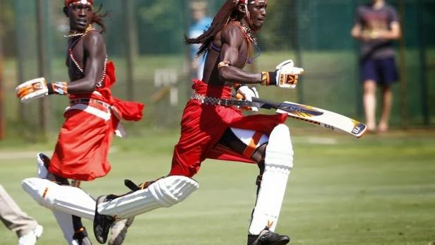 Zwei kenianische Cricketspieler in traditioneller Kleidung rennen über ein grünes Feld.