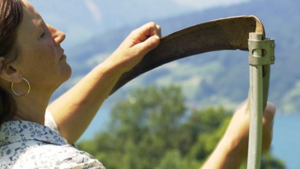 Eine Frau inspiziert eine Sense vor einer grünen Landschaft.