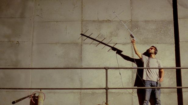 Ein Mann mit Kopfhörern und Sonnenbrille hält eine Antenne vor einer großen Wand.