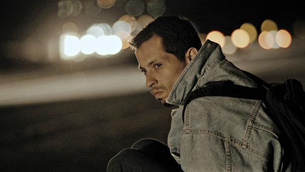 Ein Mann mit Jeansjacke und Rucksack sitzt nachts an einer Straße.