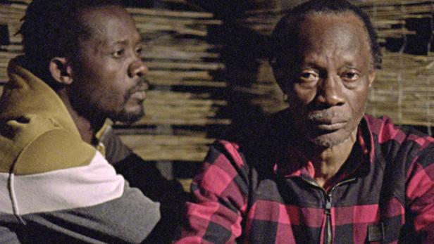 Zwei Männer sitzen nebeneinander, einer trägt ein rot-schwarz kariertes Hemd.