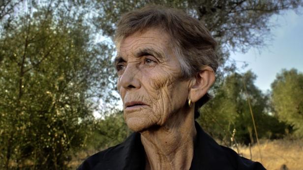 Porträt einer älteren Frau mit kurzem Haar und Ohrring vor Bäumen.