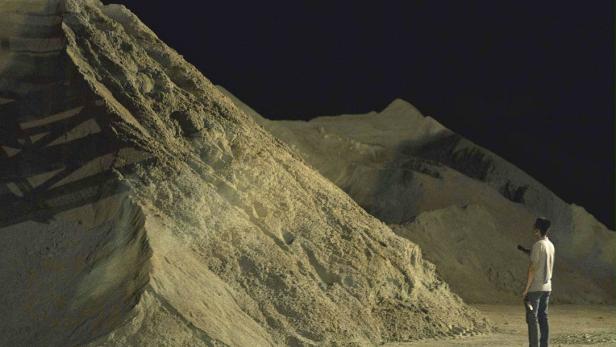 Eine Person steht vor großen Sandbergen und leuchtet mit einer Taschenlampe.
