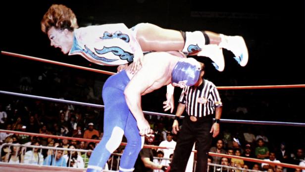 Eine Wrestlerin führt einen Move gegen einen maskierten Gegner im Ring aus, während ein Schiedsrichter zusieht.